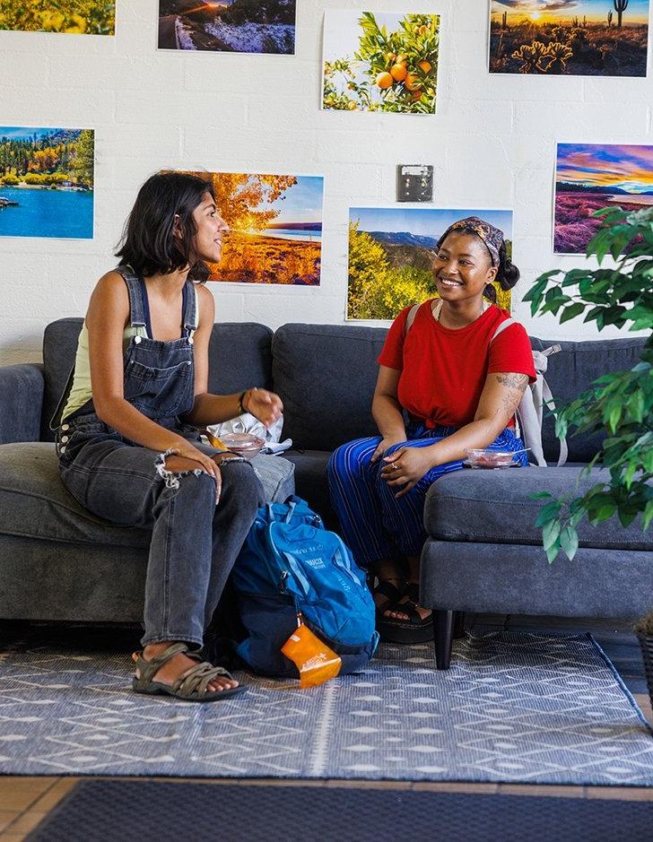 two students sit on a couch in mead lobby and chat. 